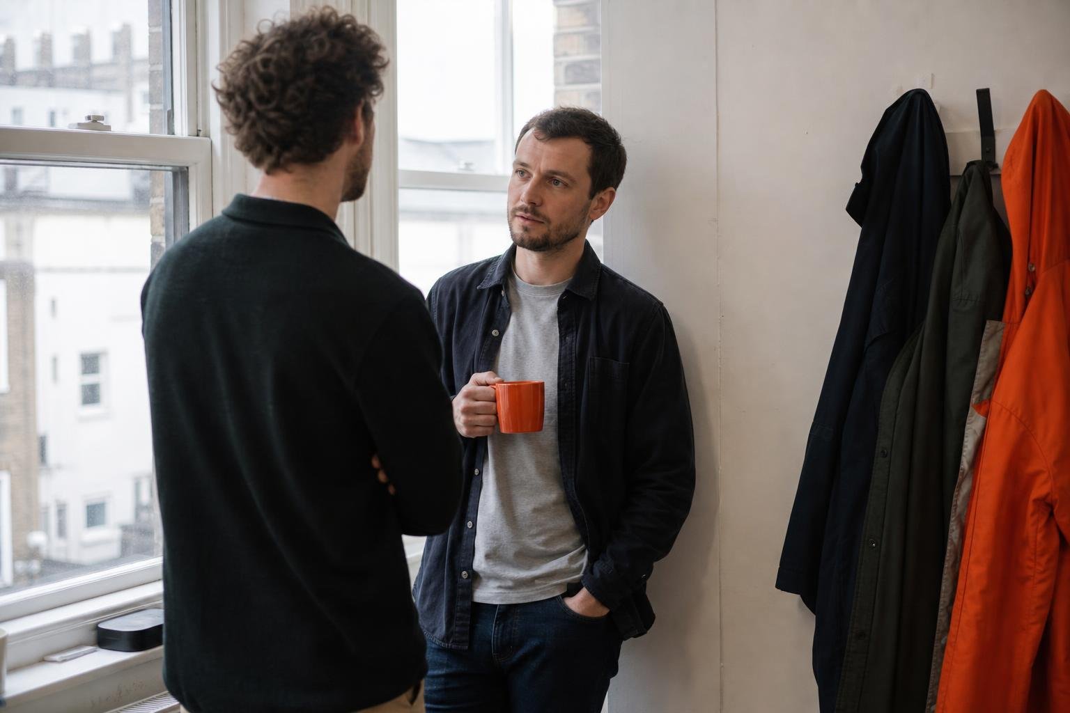 Two colleagues standing by a window in a small British office having a short conversation over a cup of tea