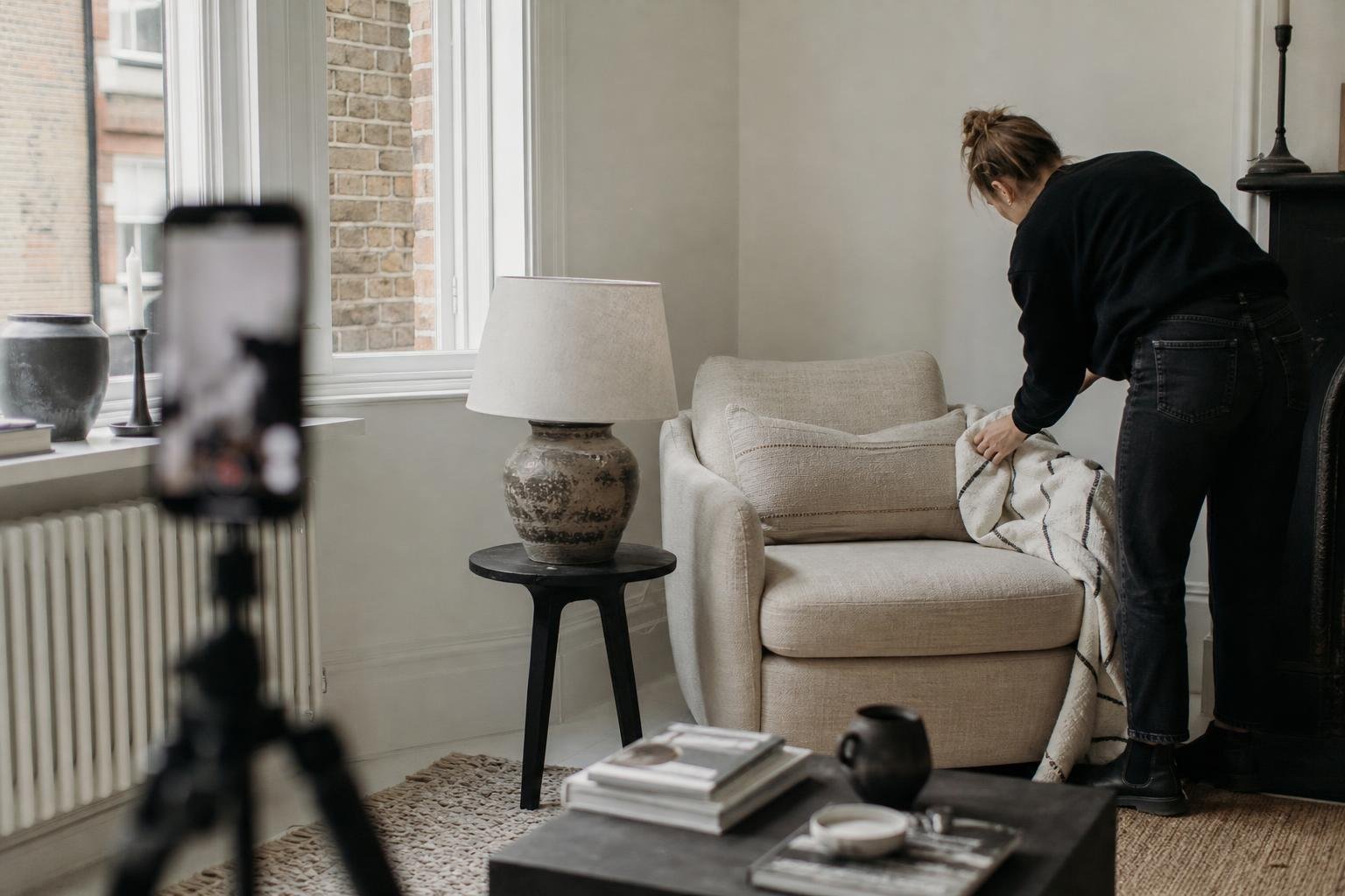 A stylist arranges a throw on a linen armchair beside a ceramic lamp, a phone on a tripod framing homeware ad creative