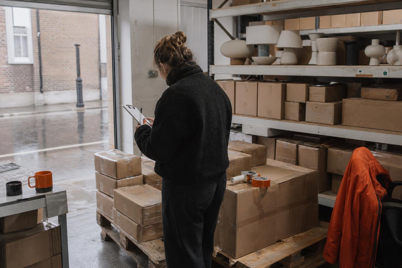 A warehouse worker checks boxed ceramic lamps against a clipboard beside an open roller door on a wet street