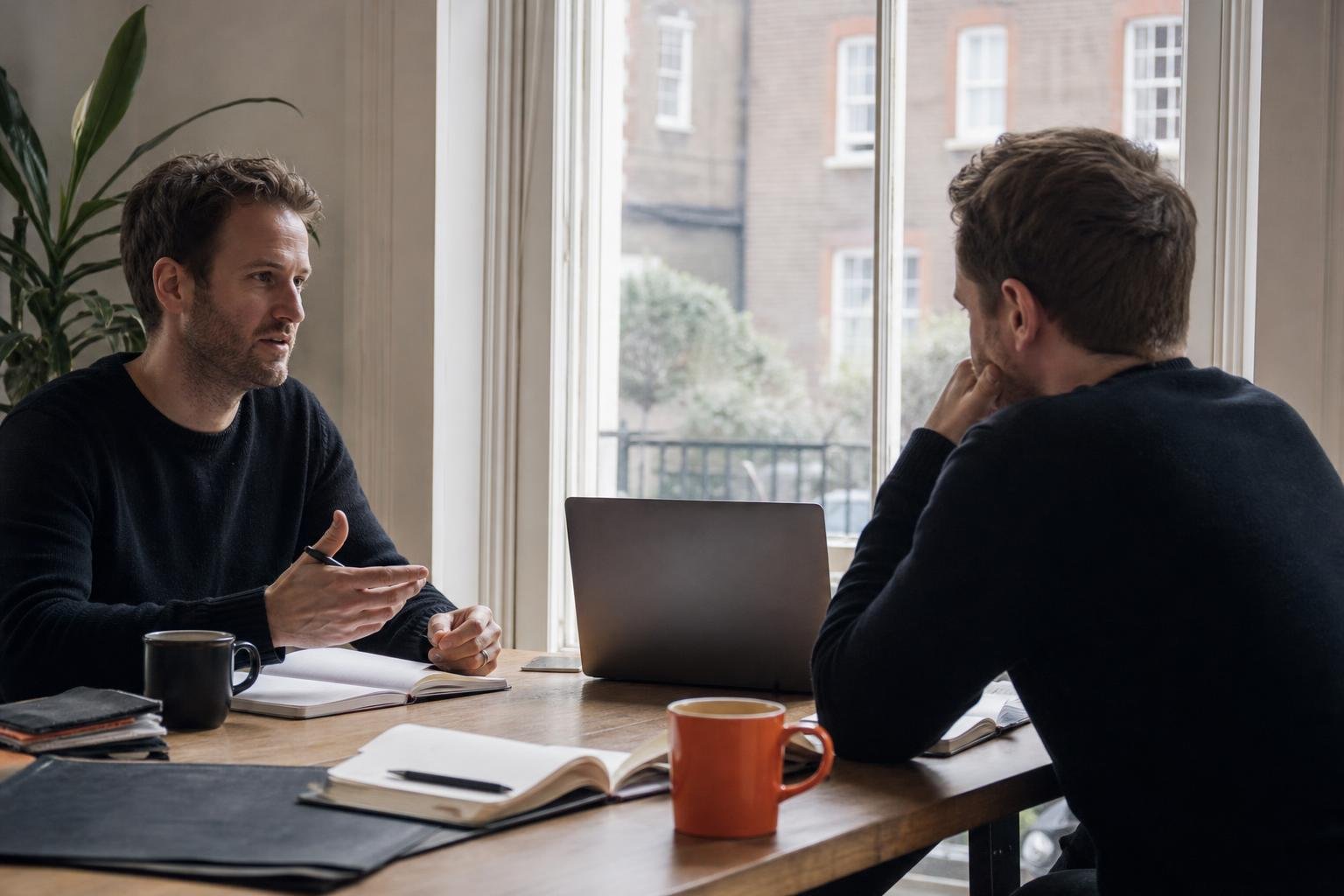 Two software co-founders talk through switching paid ads agencies at a table by a sash window