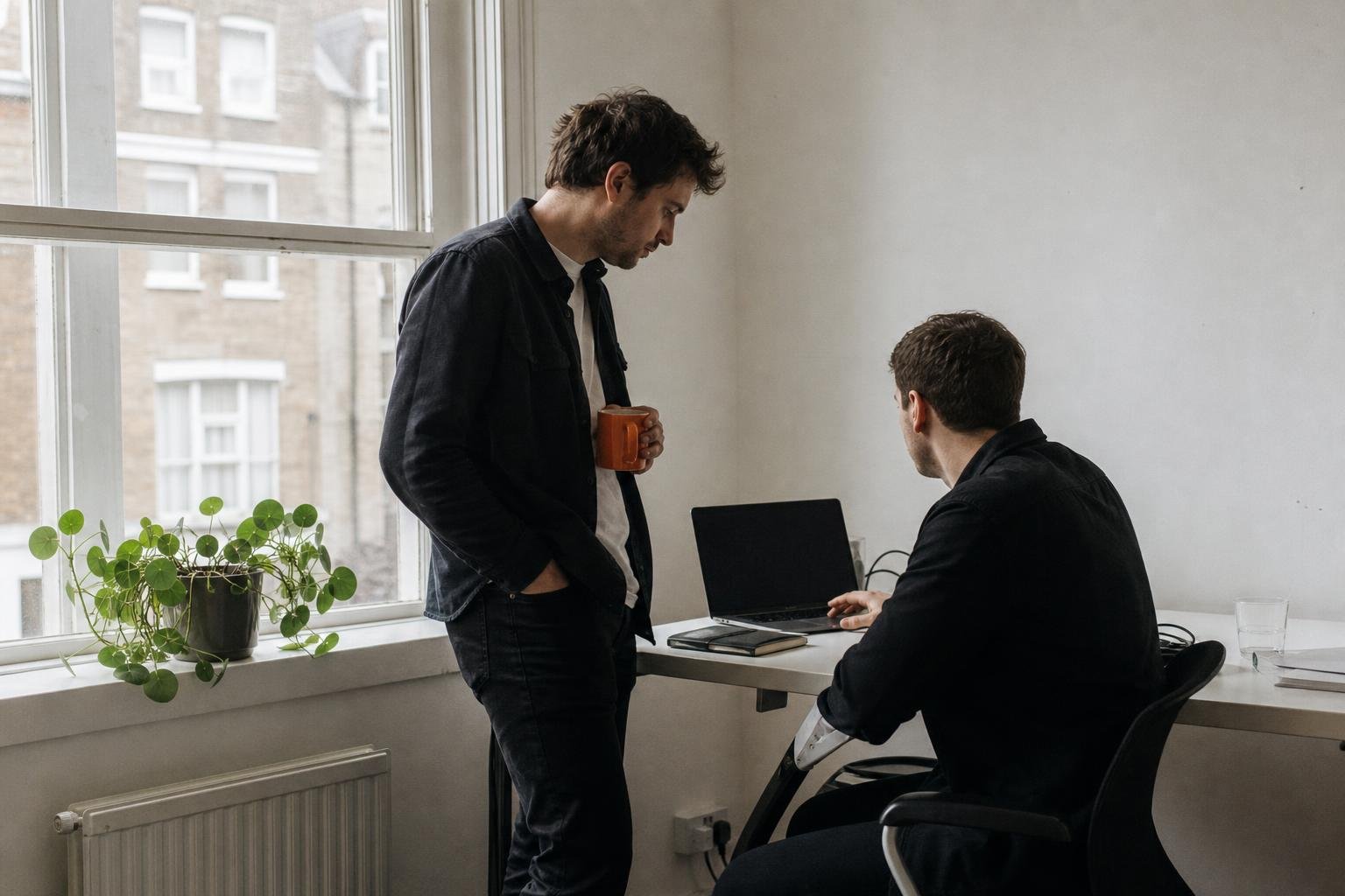 Two colleagues talking over a laptop by a tall sash window in a small British software office.