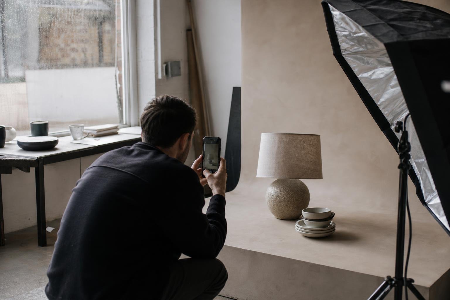 A photographer crouching to film a ceramic table lamp and stacked bowls on a plaster backdrop for a TikTok ad